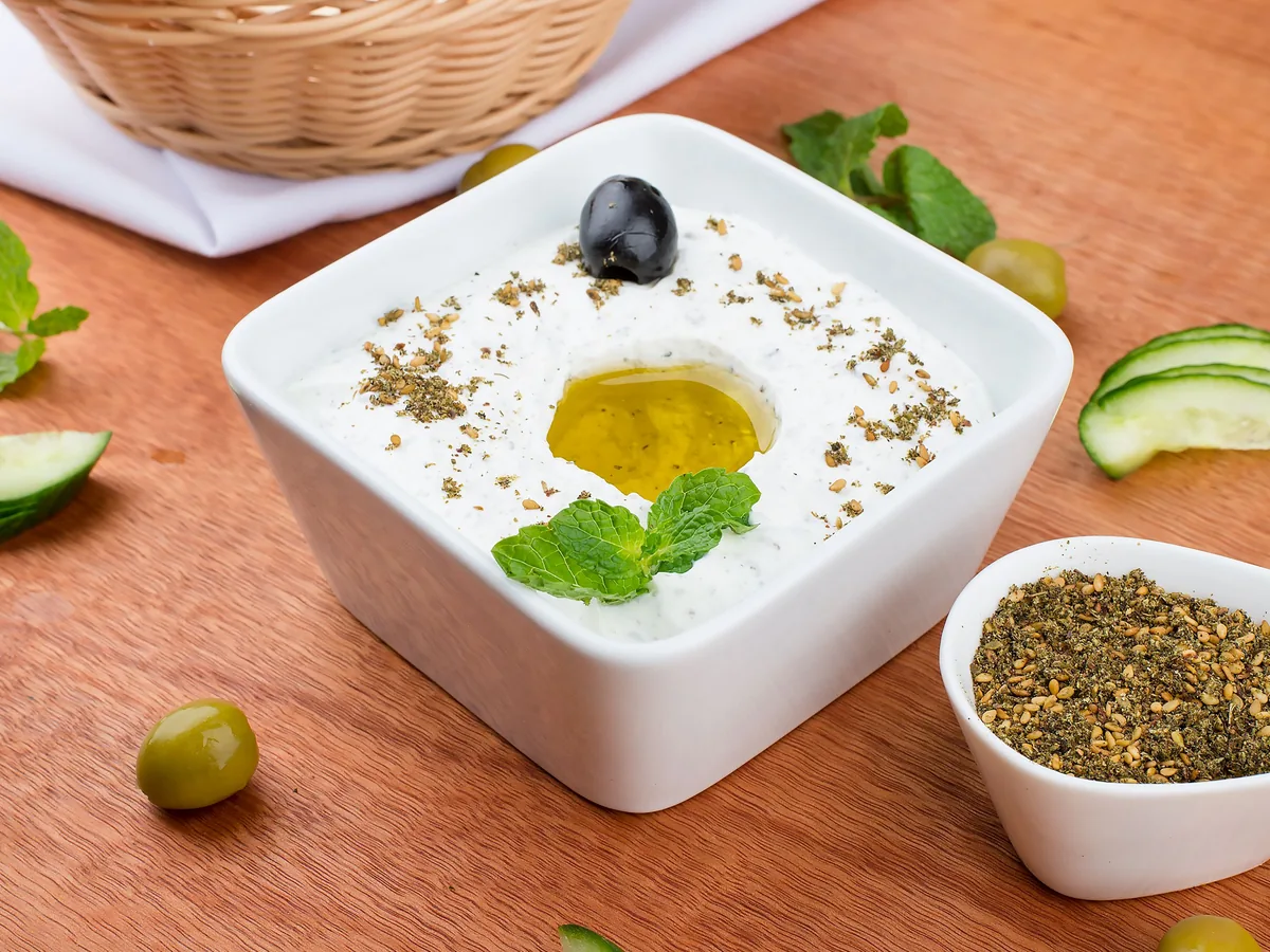 A square white bowl of creamy yogurt dip with olive oil, mint and herbs, beside olives and cucumber slices