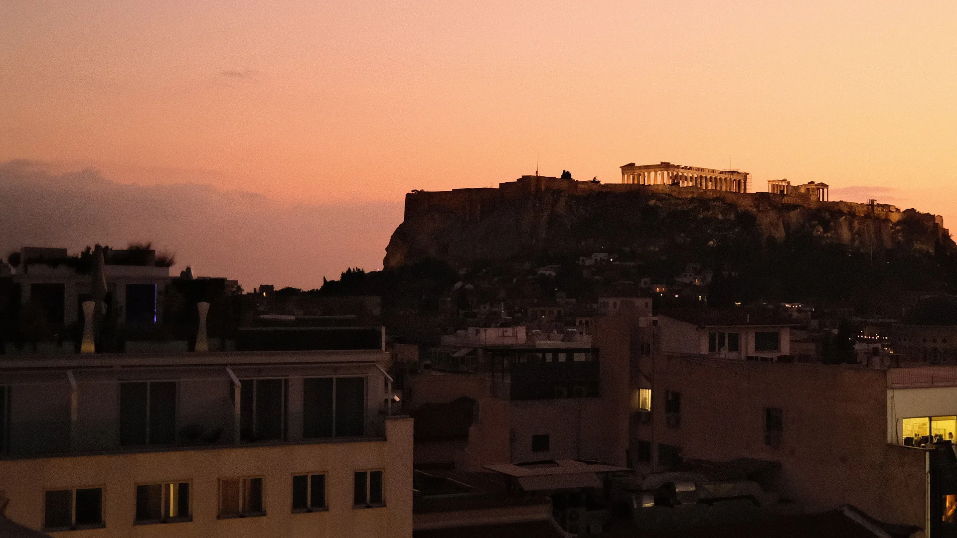 Dusk over Athens seen from a rooftop: the Parthenon floodlit on the Acropolis rock above the city's flat roofs under an orange sky