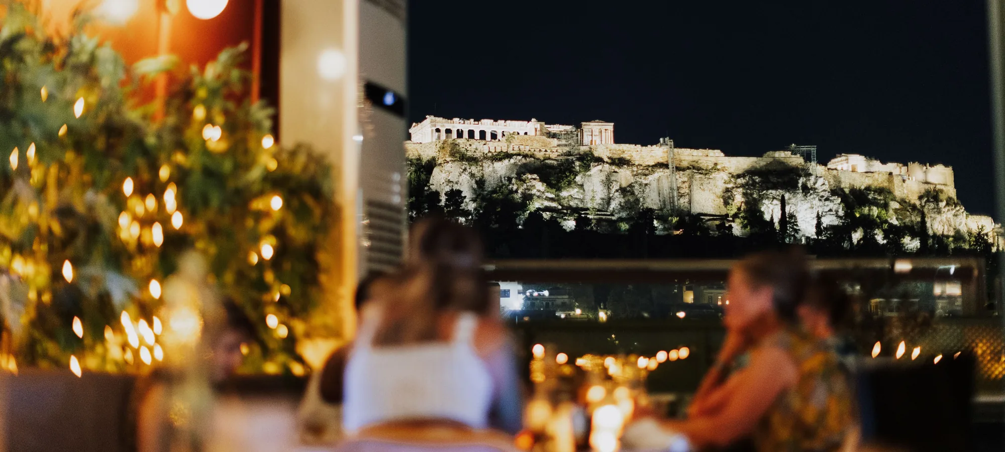 Diners at a rooftop table lit by string lights, with the floodlit Acropolis and Parthenon on the hill beyond the dark sky